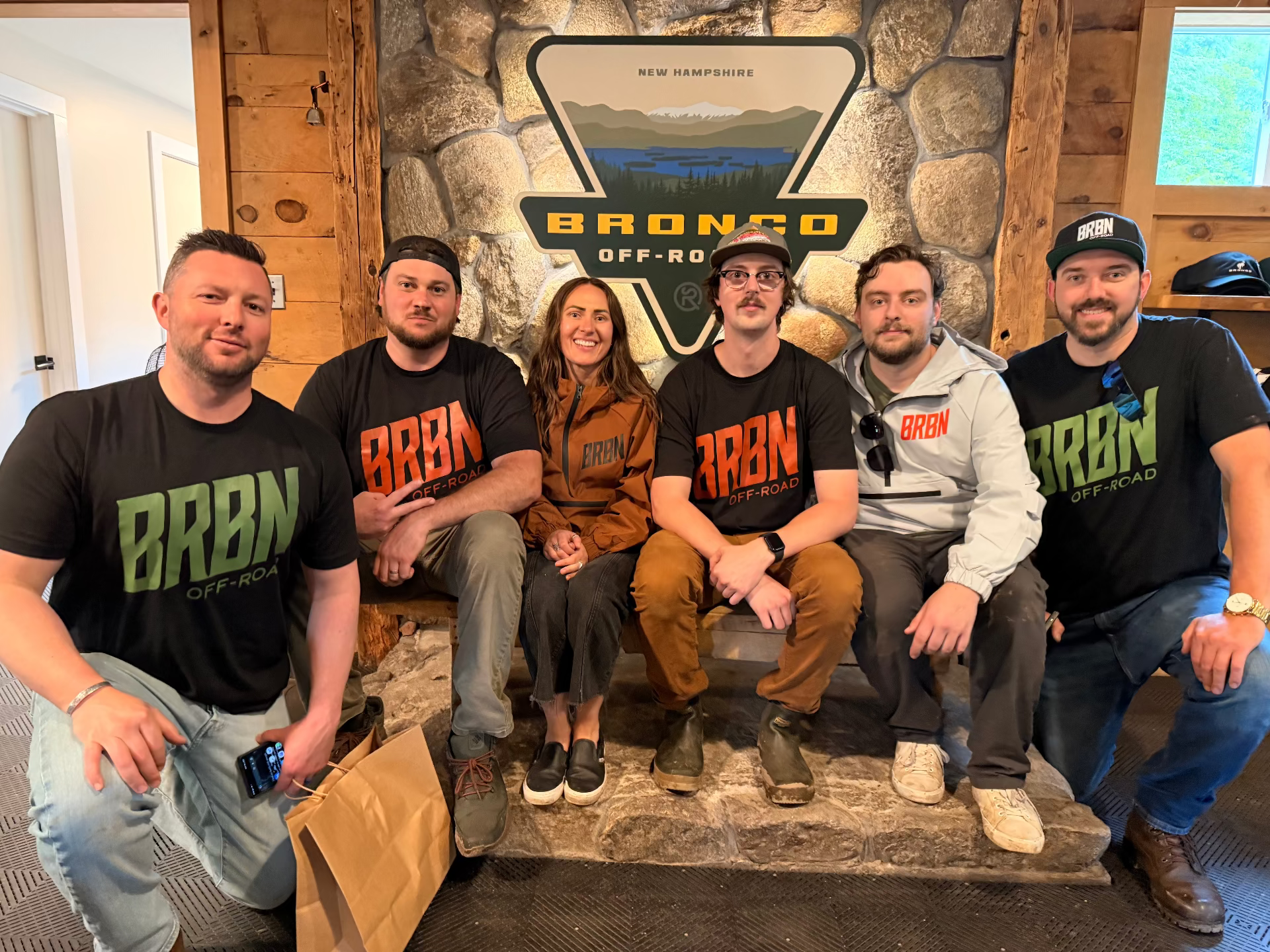 Six BRBN Off-Roaders sitting together in a rustic lodge-style room, wearing black t-shirts with 'BRBN OFF-ROAD' printed in green, orange, and white. Behind them is a large stone wall with a 'BRONCO OFF-ROAD' sign.